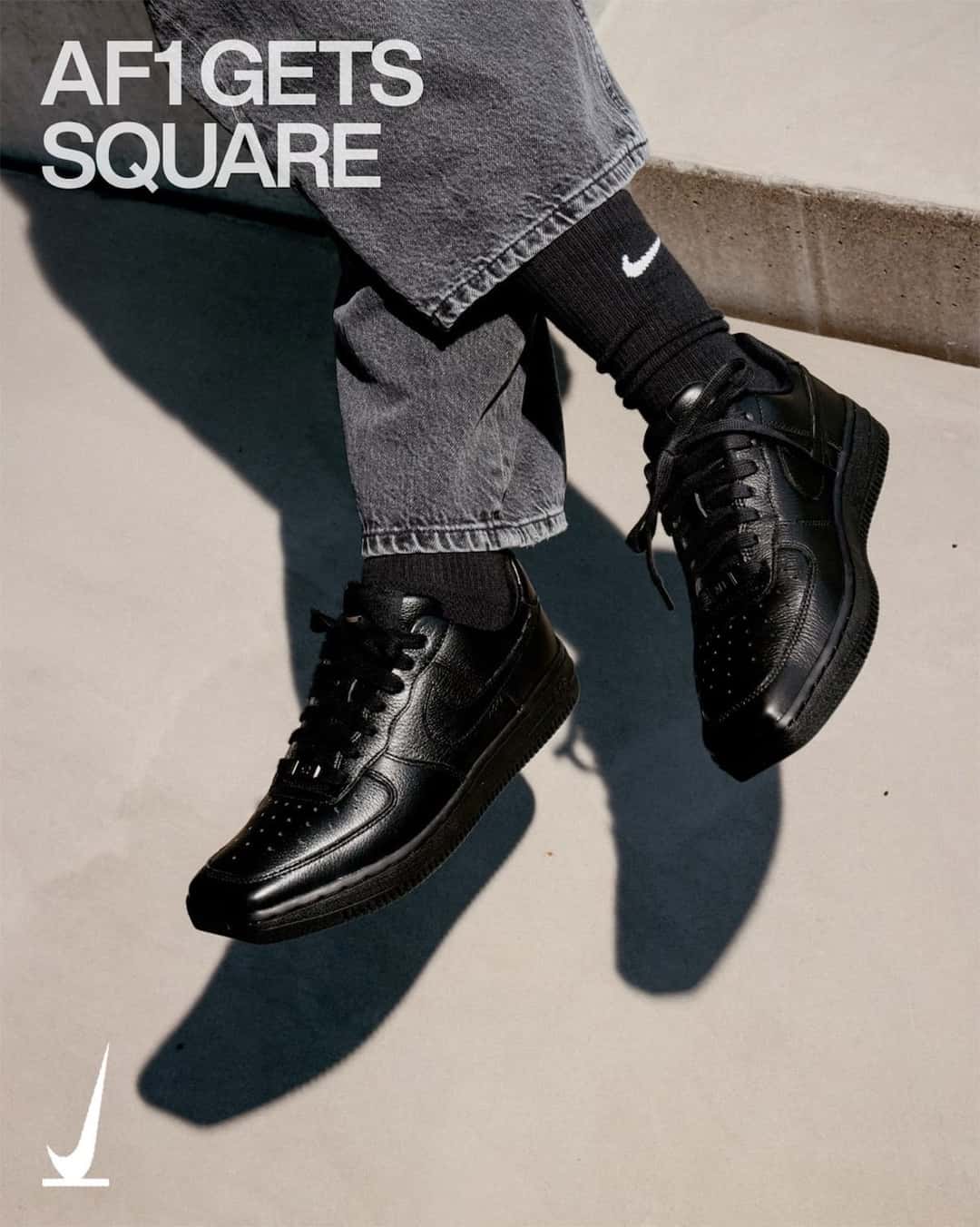Piernas cruzadas con jean gris y medias negras luciendo el Nike Air Force 1 SquAir Toe en cuero negro entero, con la puntera de forma cuadrada, en la imagen oficial de SNKRS con la frase AF1 Gets Square