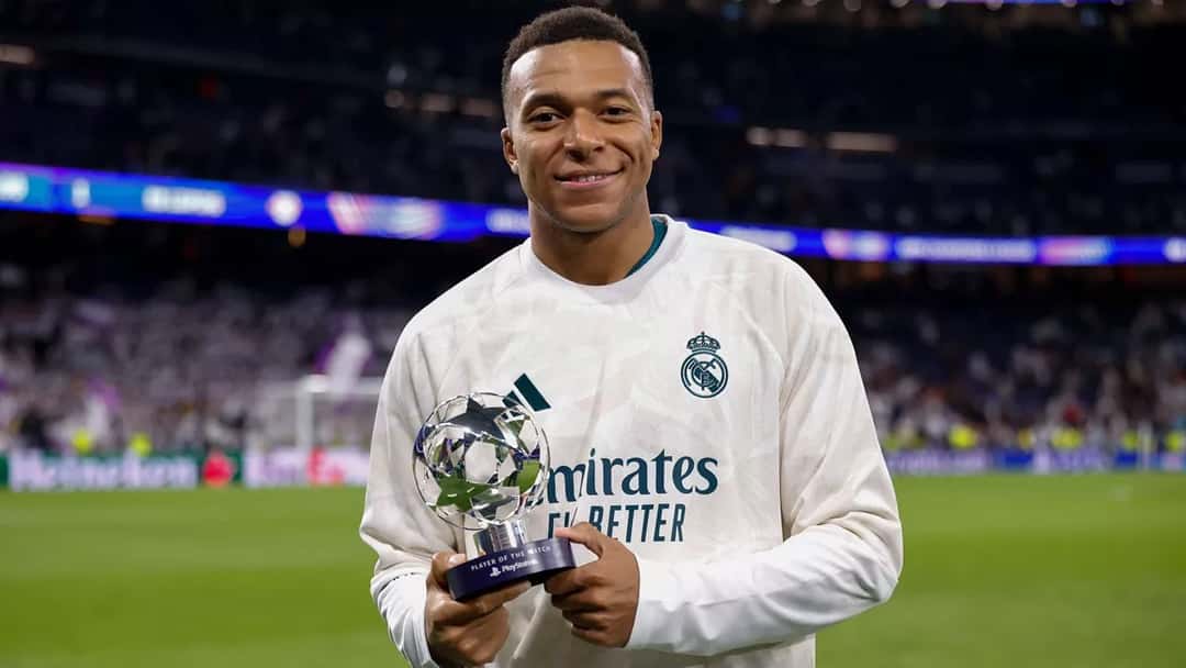 Kylian Mbappé, con buzo blanco del Real Madrid, sonríe con el trofeo de jugador del partido en la mano en el césped del Bernabéu (foto: Real Madrid)