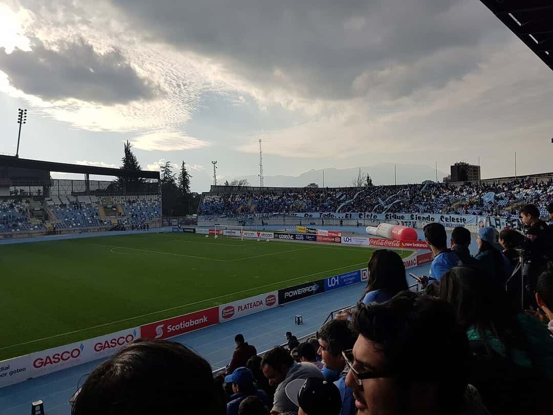 Las tribunas del estadio El Teniente, en Rancagua, con hinchas en primer plano y la cordillera al fondo; foto de archivo de 2017 (foto: Aeveraal, Wikimedia Commons, licencia CC BY-SA 4.0)