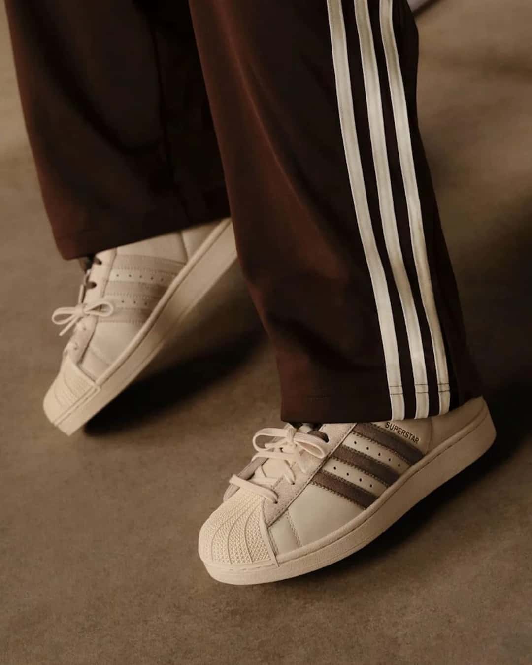 Pies con el Molly-Mae x adidas Superstar Latte color crema con tiras marrones, junto a un pantalón marrón de adidas con tres tiras blancas