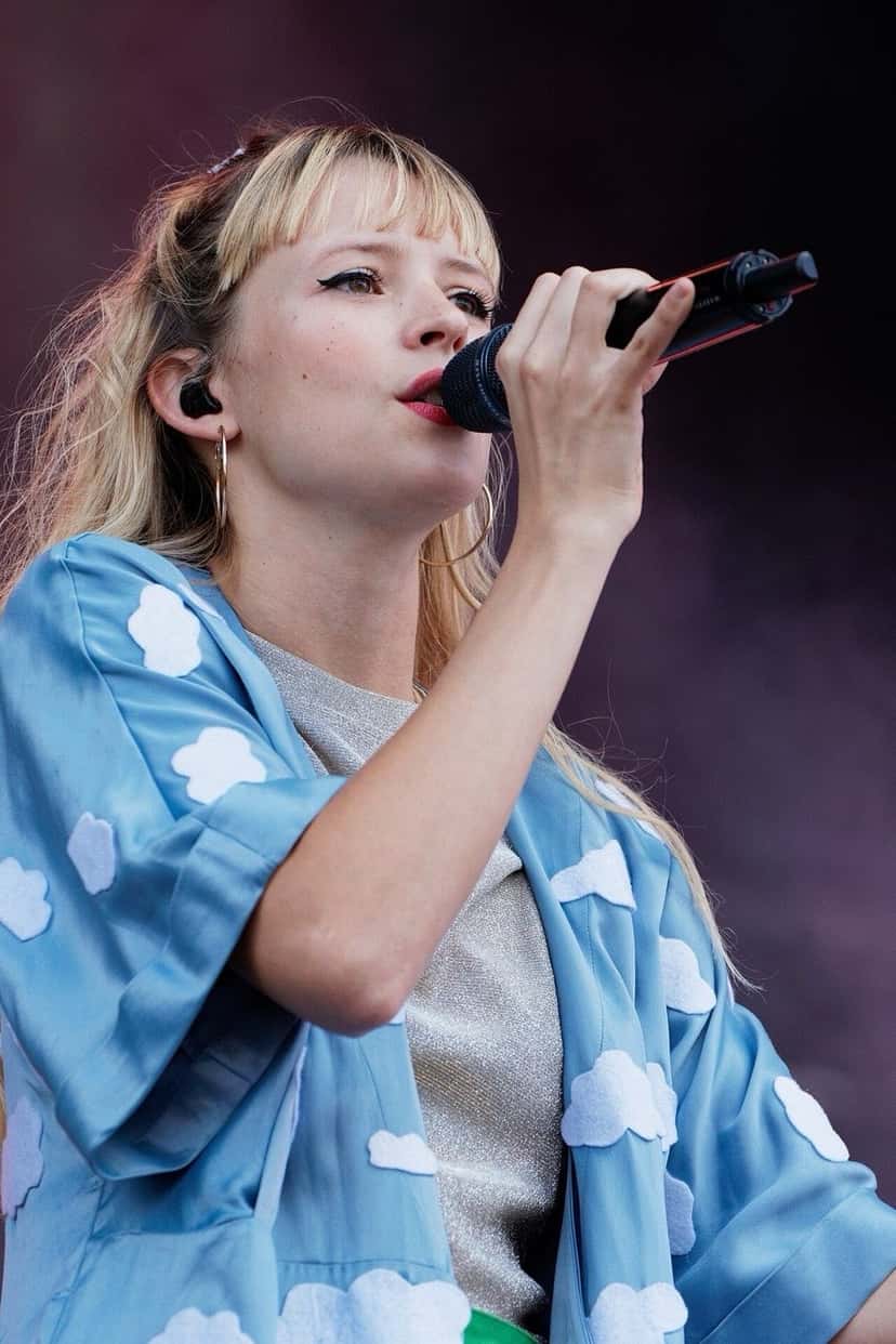 Angèle canta con un micrófono negro en el festival Vieilles Charrues de 2018, con una camisa celeste con nubes blancas y aros grandes (foto de archivo). Foto: Thesupermat, CC BY-SA 4.0, vía Wikimedia Commons