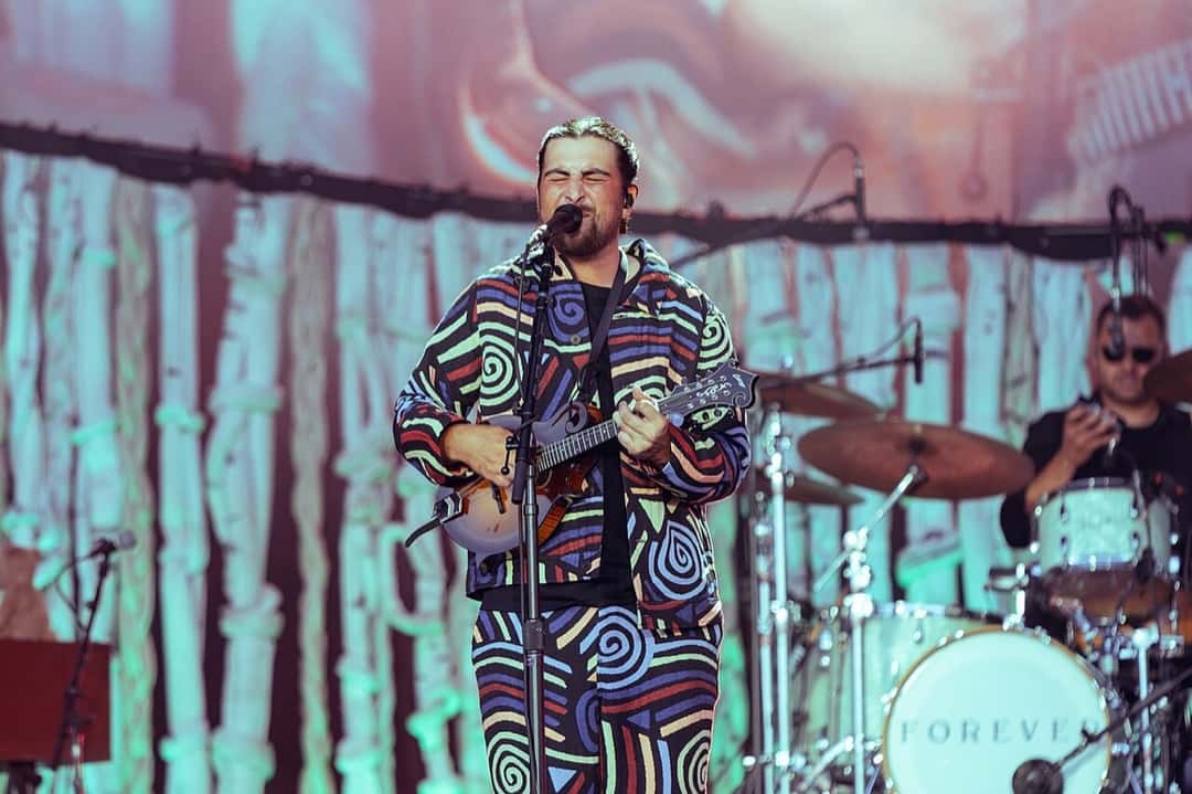 Noah Kahan canta y toca una mandolina en el Glastonbury Festival de 2025, con el mismo traje de espirales de colores y un baterista detrás (foto de archivo). Foto: Raph_PH, CC BY 4.0, vía Wikimedia Commons