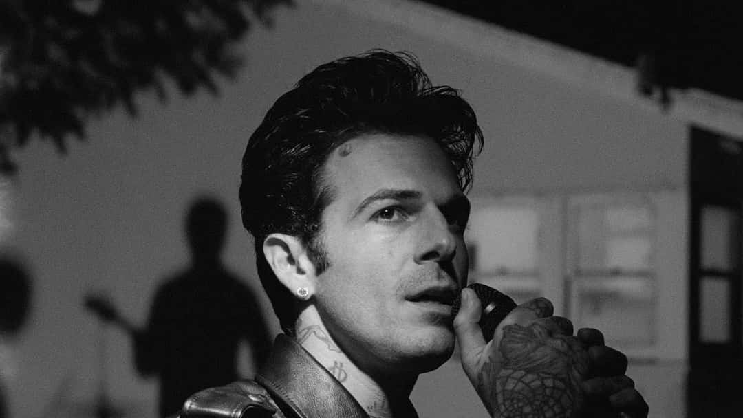 Jesse Rutherford, cantante de The Neighbourhood, con campera oscura y tatuajes en la mano, sostiene un micrófono frente a una casa, en una imagen en blanco y negro del video oficial de «Private»
