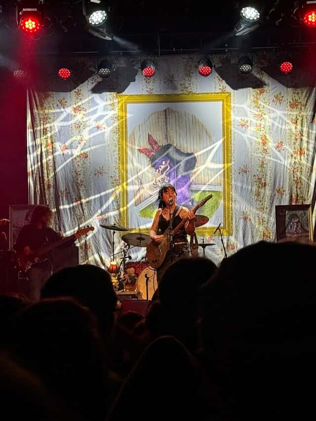 Karly Hartzman canta y toca la guitarra en el centro del escenario con Wednesday, frente a un telón floreado con un cuadro y una telaraña de luces, en un show en el First Avenue de Minneapolis, en marzo de 2026 (foto de archivo). Foto: Marchiviste, CC BY-SA 4.0, vía Wikimedia Commons