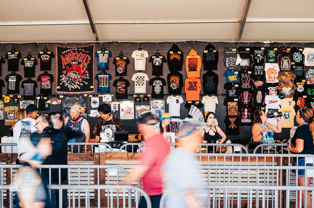 Un puesto de remeras de bandas en Welcome to Rockville 2024: paredes cubiertas de remeras negras y estampadas, con gente comprando detrás de una valla y otros caminando borrosos en primer plano (foto de archivo). Imagen: Welcome to Rockville