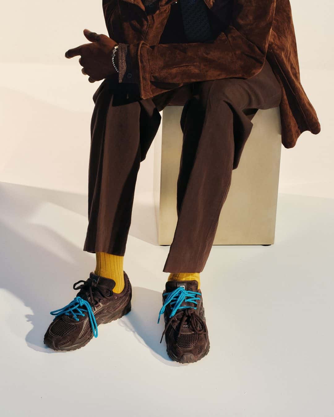 Persona sentada con las BILLY'S x New Balance 204L marrones y cordones turquesa, medias amarillas y saco de gamuza