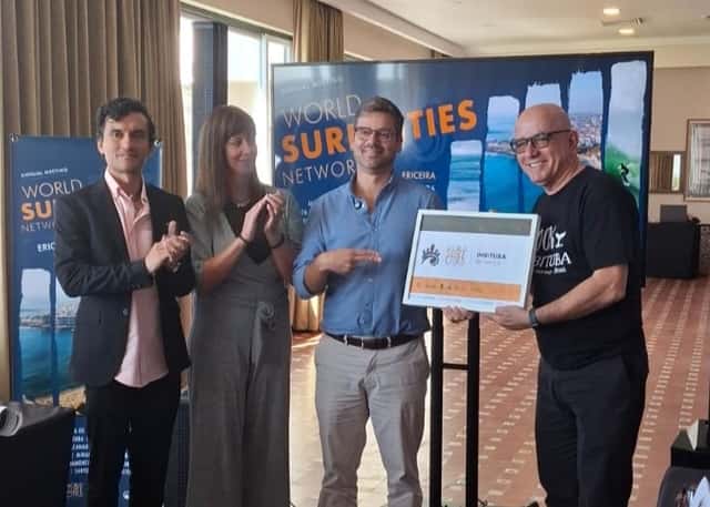 Cuatro personas en la asamblea de la World Surf Cities Network en Ericeira; un representante de Imbituba, con remera negra, recibe el certificado de la ciudad