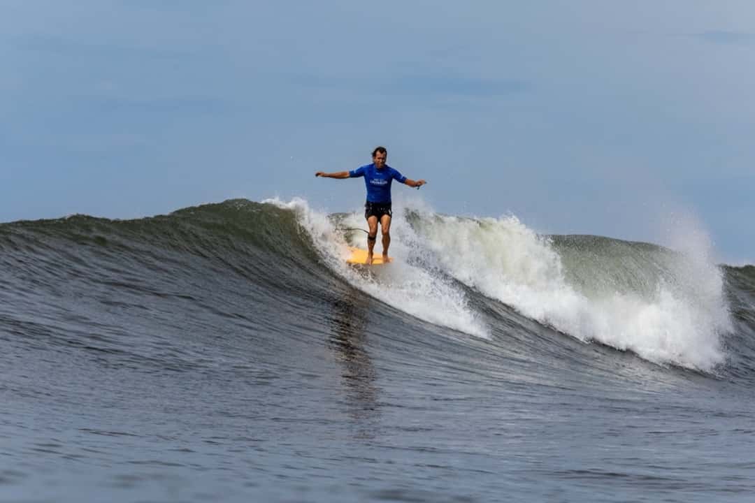 Tony Silvagni, con lycra azul, camina sobre un longboard amarillo con los brazos abiertos en una ola de El Sunzal