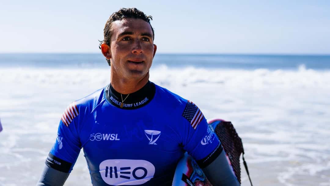 Griffin Colapinto, con la lycra azul del MEO Rip Curl Pro Portugal y la bandera de Estados Unidos, mira a un costado con el mar de fondo en Supertubos