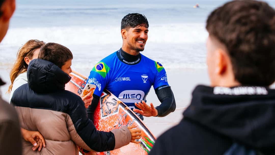 Gabriel Medina, con la lycra azul del MEO Rip Curl Pro Portugal y la bandera de Brasil, sonríe con su tabla naranja mientras lo saludan en la orilla de Supertubos