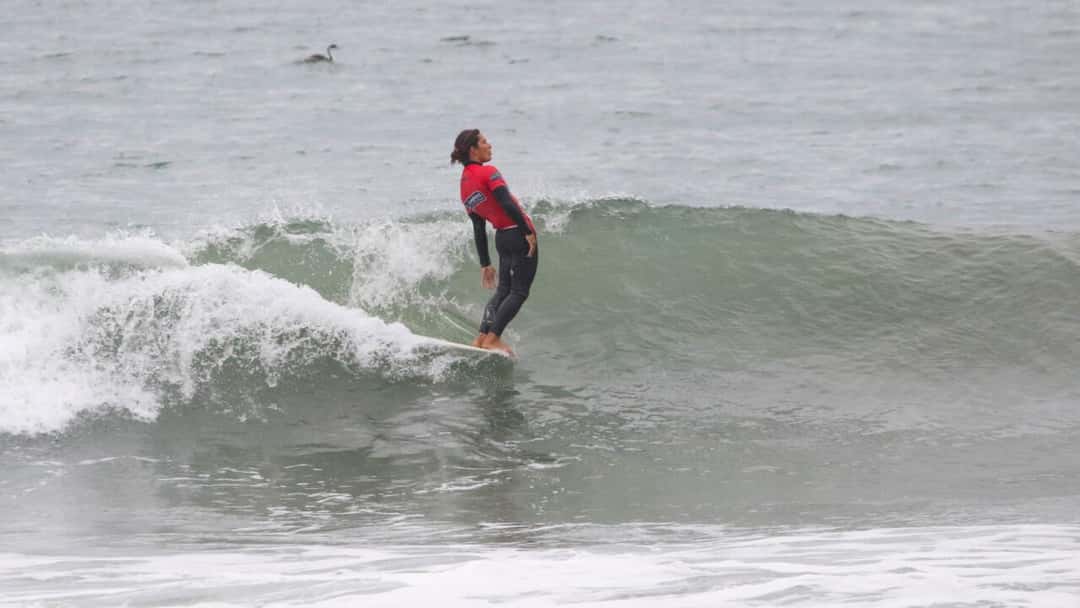 Jonathan Melendres, con lycra roja y traje negro, se inclina hacia atrás parado sobre su longboard mientras la ola se cierra a su espalda en Pismo Beach