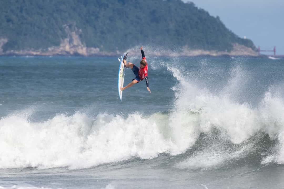 Ryan Kainalo vuela en un aéreo sobre una ola en Maresias, con el morro verde de la costa paulista de fondo