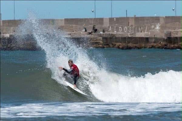 Un surfista con lycra roja hace una maniobra cerrada sobre la pared de la ola, con un paredón de piedra detrás