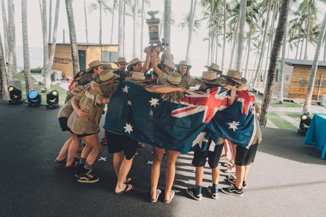 El equipo junior de Australia abrazado bajo una bandera australiana levanta el trofeo por equipos entre palmeras en El Salvador (foto de archivo)
