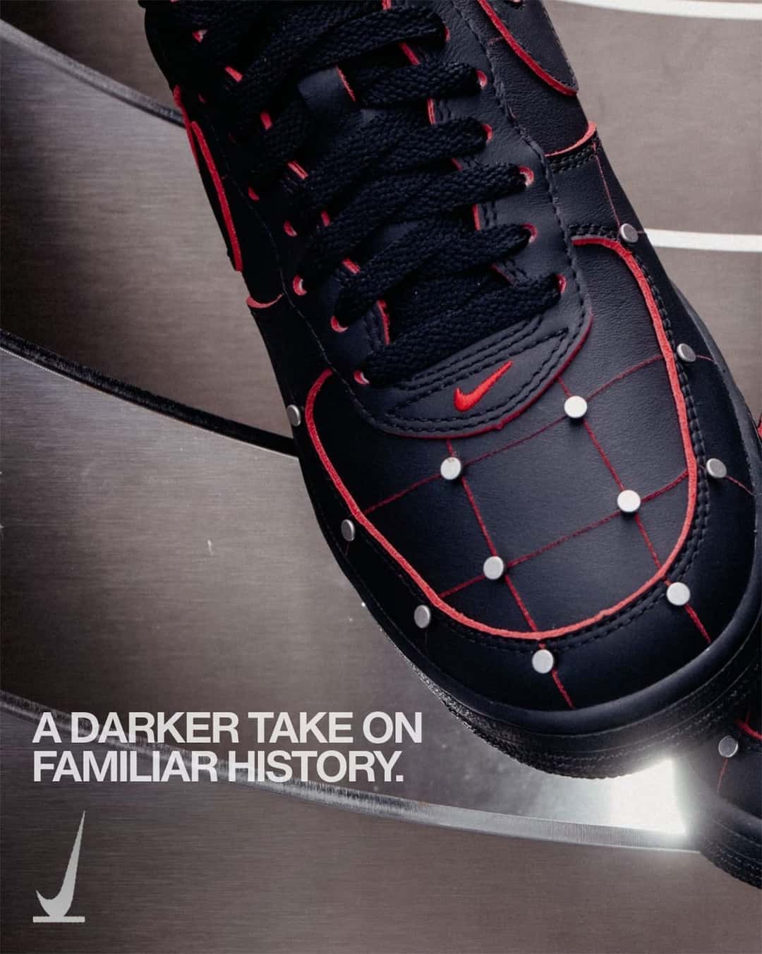 Primer plano de la puntera del Nike Air Force 1 '01 Nightmare en cuero negro, con costuras rojas que forman una cuadrícula, tachas plateadas en cada cruce, cordones negros, ojales con borde rojo y swoosh rojo chico, sobre una superficie metálica curva