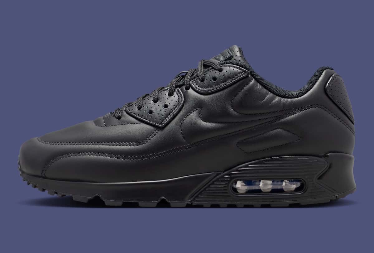 Nike Air Max 90 SP Black Leather de perfil sobre fondo azul grisáceo, toda de cuero negro con el Swoosh y el mudguard marcados con costuras, cordones negros y la cámara Air visible en el talón