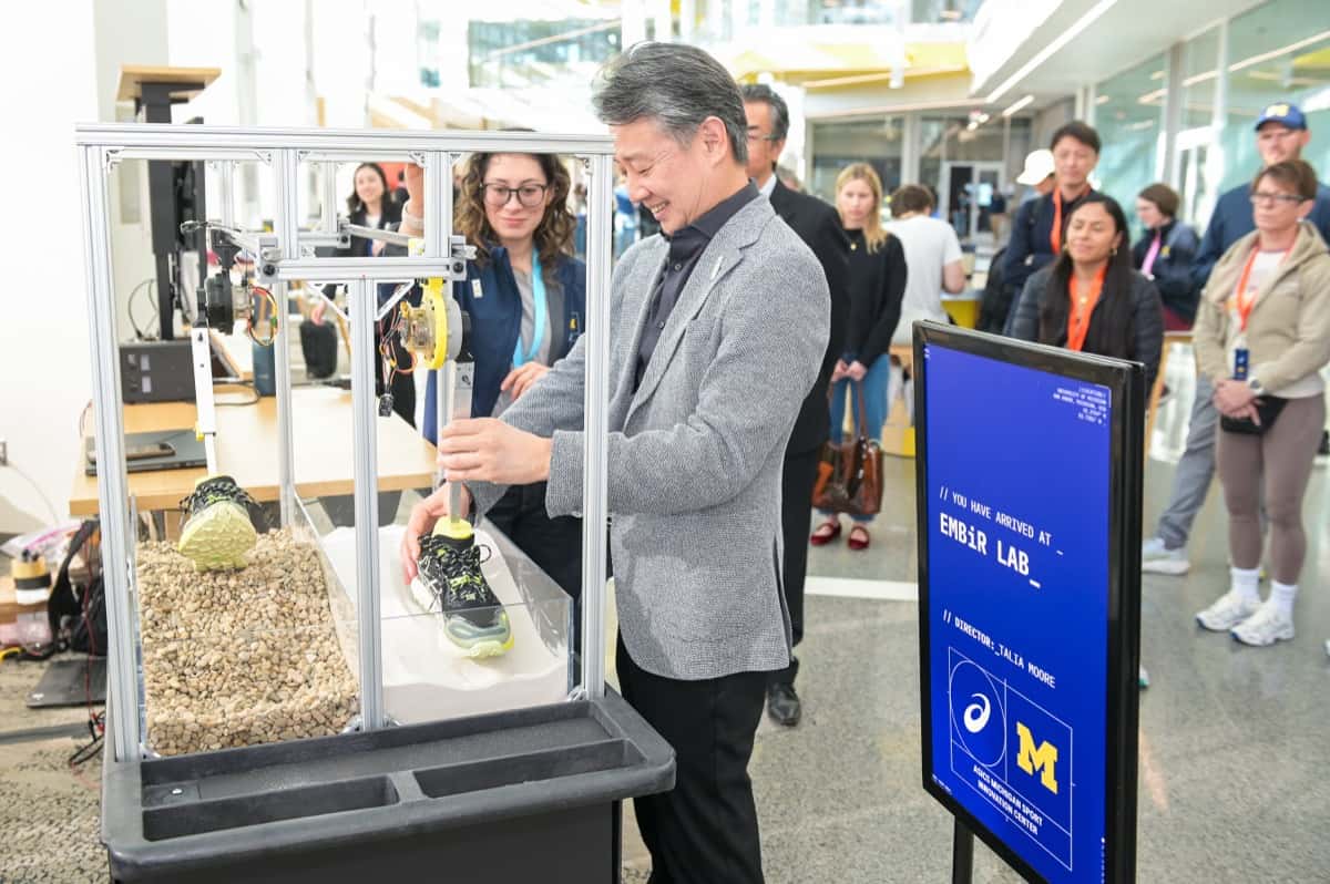 Visitantes en la inauguración del AMSI Lab de ASICS en la Universidad de Michigan, junto a una estación de pruebas con una zapatilla y un cartel con los logos de ASICS y Michigan
