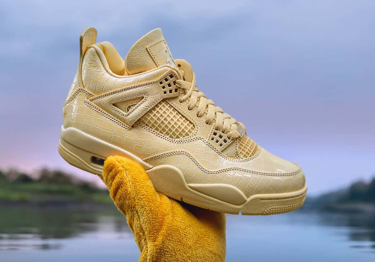 J Balvin x Air Jordan 4 Amazonas en cuero amarillo con textura de cocodrilo, sostenida frente a un río al atardecer