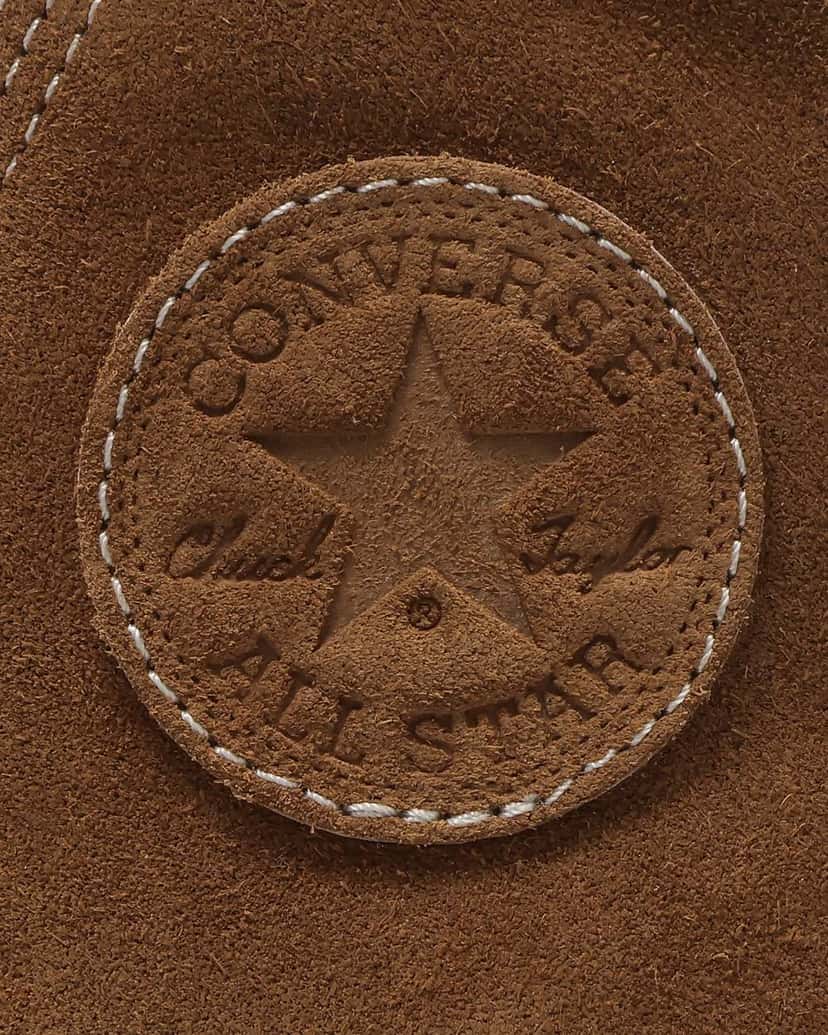 Primer plano del parche del tobillo de la versión tan: un círculo de gamuza marrón con la estrella y las palabras Converse, Chuck Taylor y All Star grabadas en relieve, rodeado por una costura en hilo claro