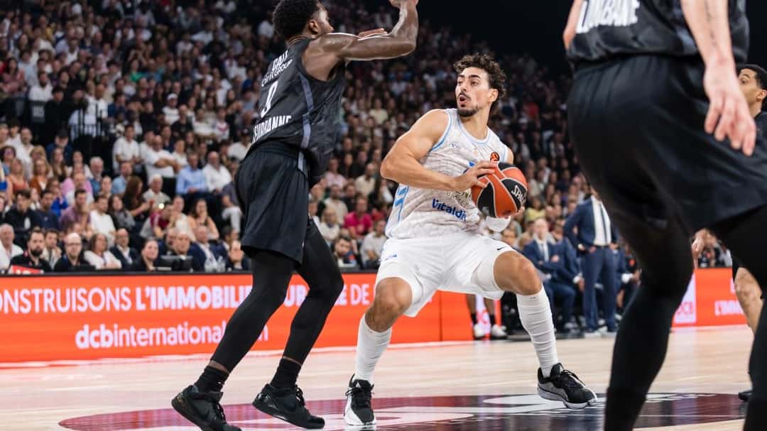 Un jugador del Valencia Basket, con la camiseta blanca, protege la pelota ante la marca de un jugador del LDLC ASVEL de camiseta negra, con la tribuna llena detrás