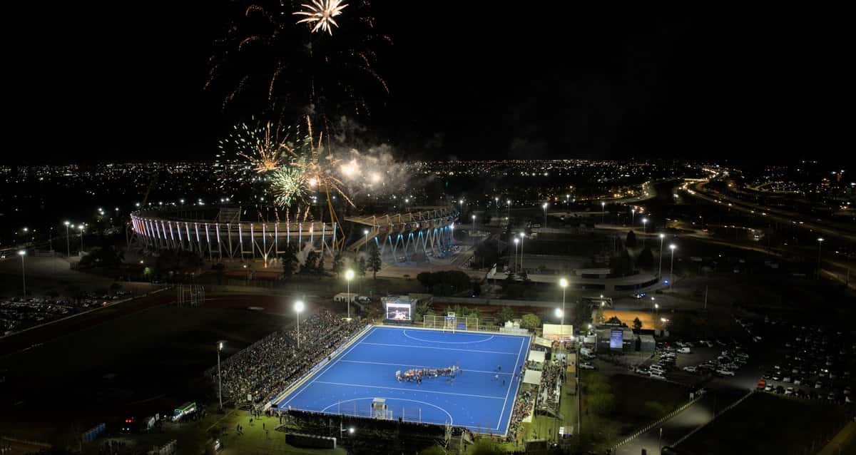Vista aérea nocturna de la cancha azul del estadio de hockey Soledad García, con las tribunas llenas y fuegos artificiales sobre el Estadio Kempes