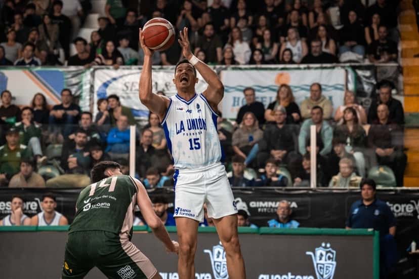 Un jugador de La Unión de Formosa con la camiseta blanca 15 lanza en suspensión ante la marca de un jugador de Gimnasia con la camiseta verde 14, con la tribuna llena de fondo