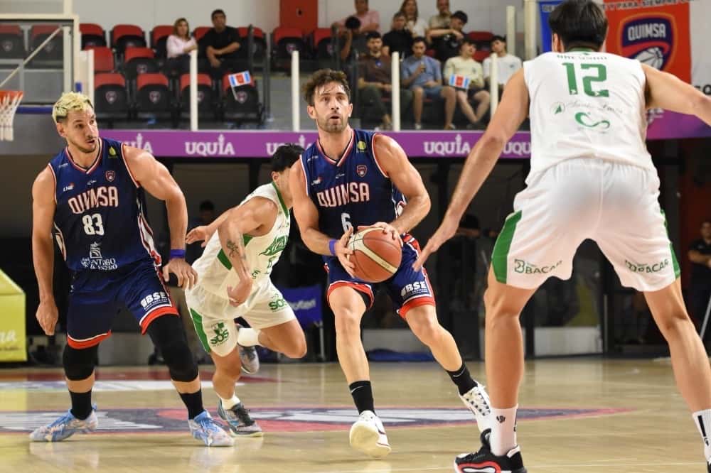 Un jugador de Quimsa con la camiseta azul 6 maneja la pelota ante la marca de jugadores de Atenas con camiseta blanca y verde, en el Estadio Ciudad de Santiago del Estero