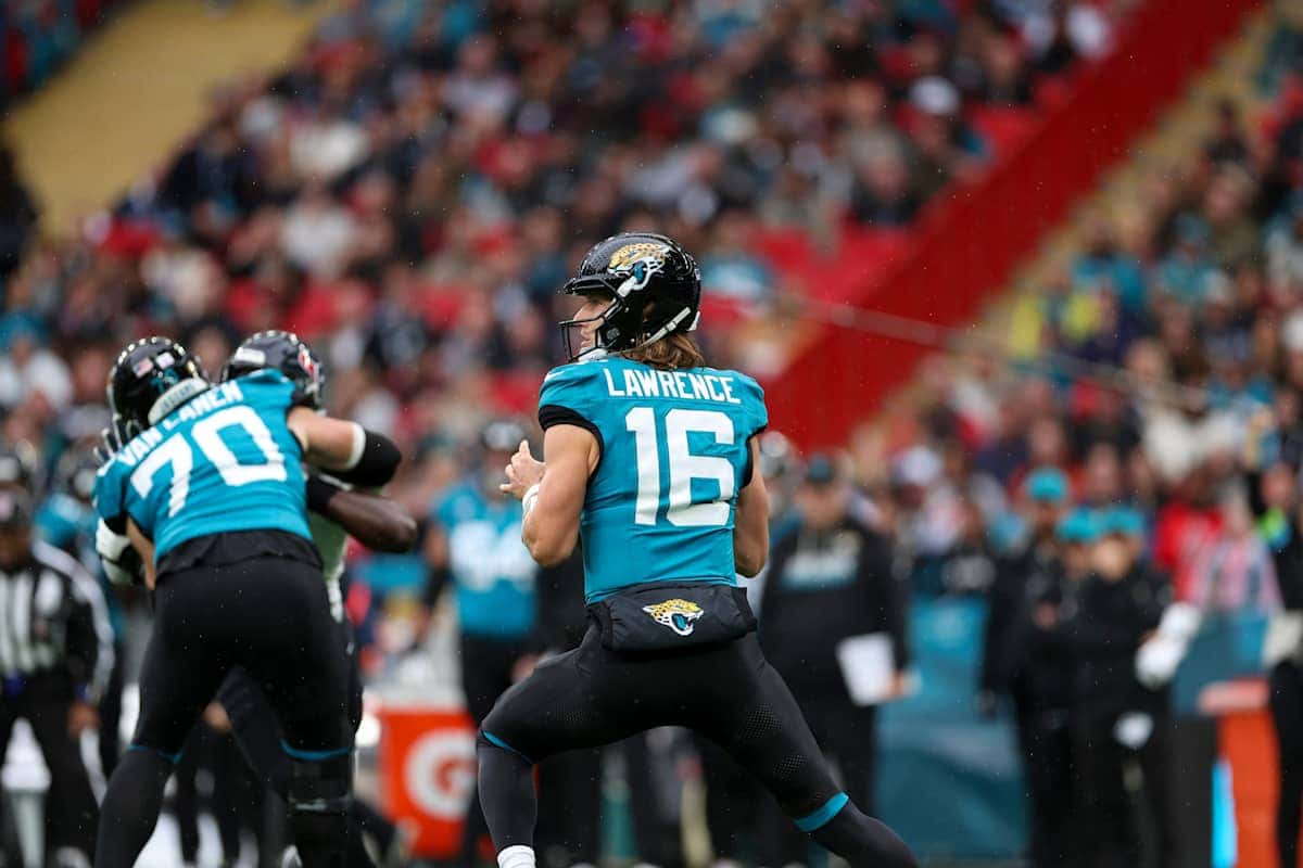 Trevor Lawrence, con la camiseta turquesa número 16 de los Jaguars, prepara un pase detrás de su línea, con el número 70 bloqueando a su izquierda y la tribuna de Wembley detrás