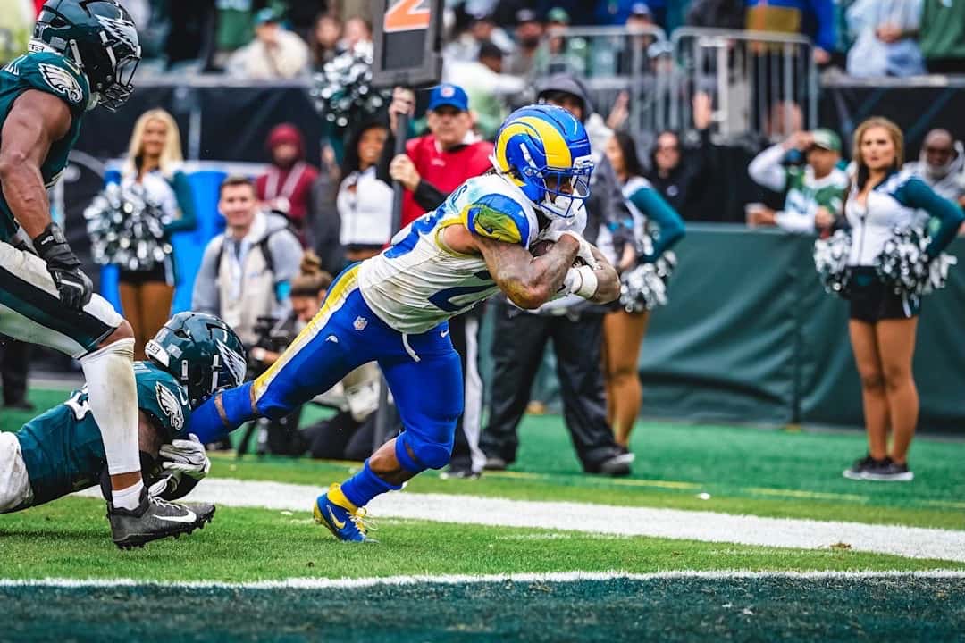 Un corredor de los Rams, de camiseta blanca y casco azul y amarillo, se lanza hacia la zona de anotación mientras un defensor de los Eagles lo agarra desde el piso en el Lincoln Financial Field