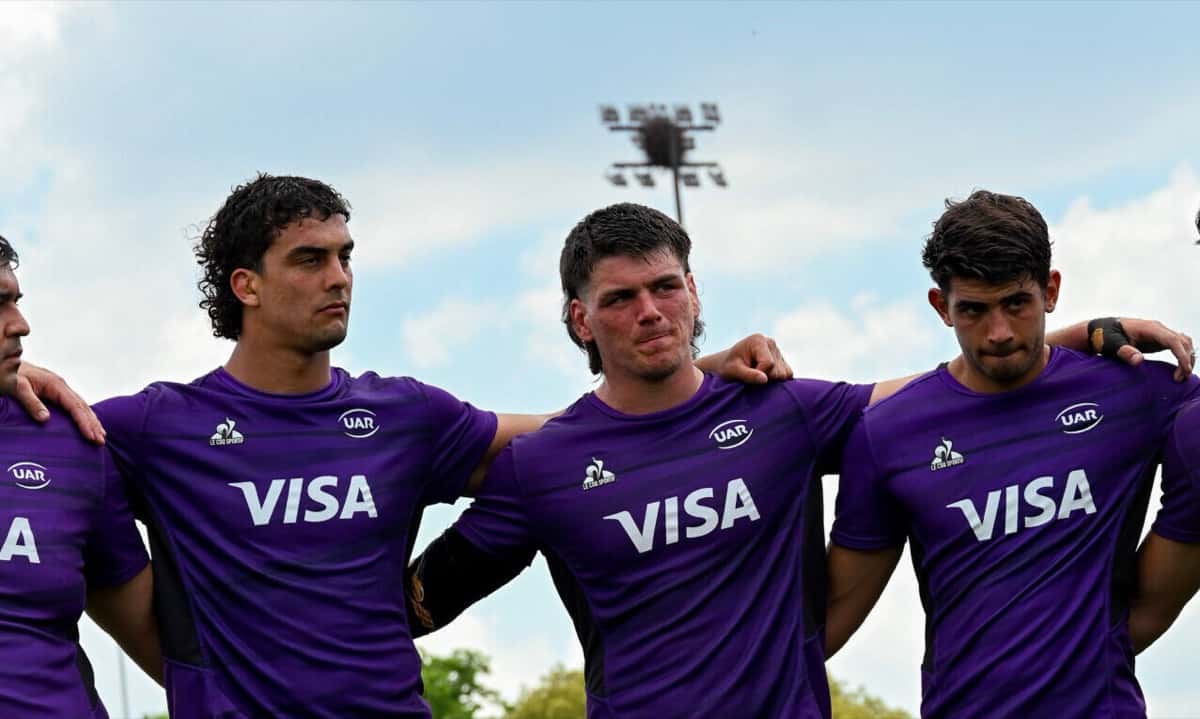 Tres jugadores de Argentina XV, con la remera violeta de entrenamiento de la UAR, abrazados en una cancha bajo el cielo nublado