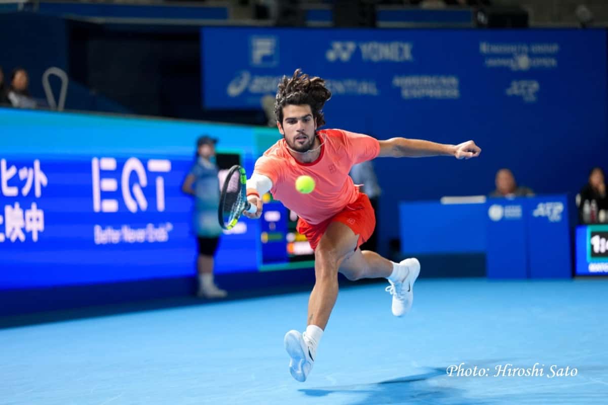 Carlos Alcaraz, con remera coral y short rojo, se estira para pegar un revés frente a los carteles azules del Japan Open en el Ariake Coliseum