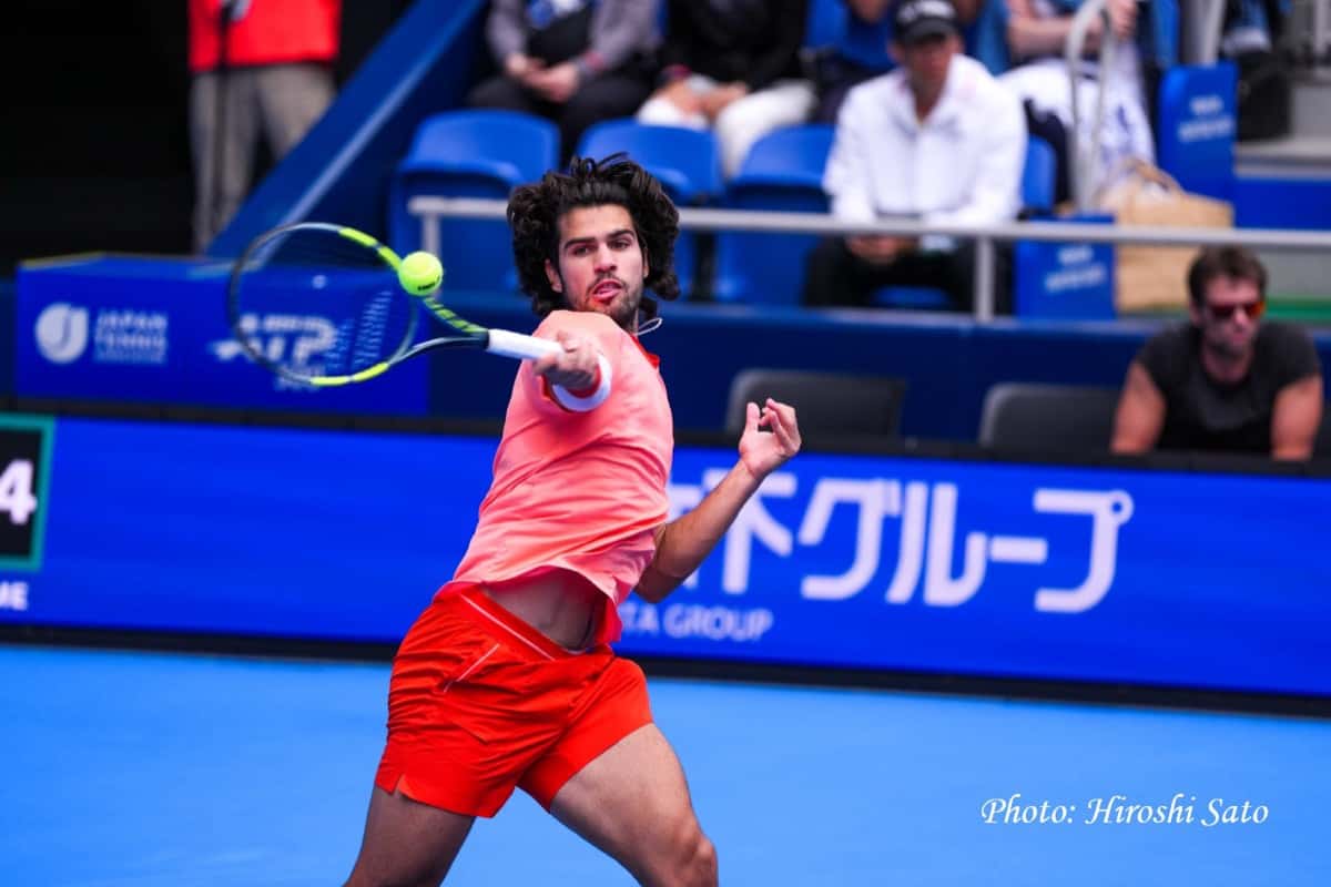 Carlos Alcaraz, con remera coral y short rojo, pega una derecha frente al cartel azul del Japan Open en el Ariake Coliseum