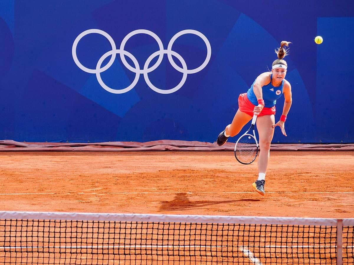 Foto de archivo: Karolína Muchová, con remera azul y pollera roja, saca en una cancha de polvo de ladrillo con los anillos olímpicos de fondo