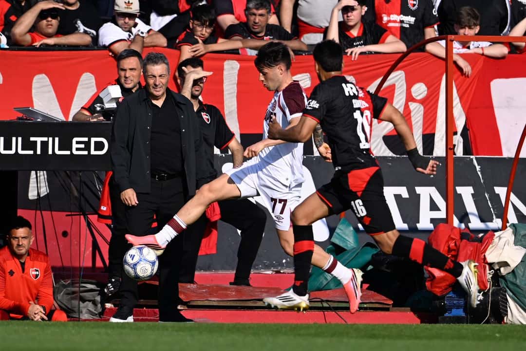 Un jugador de Lanús, con camiseta blanca y el número 77, controla la pelota junto a la línea de banda perseguido por un futbolista de Newell's en el Coloso Marcelo Bielsa
