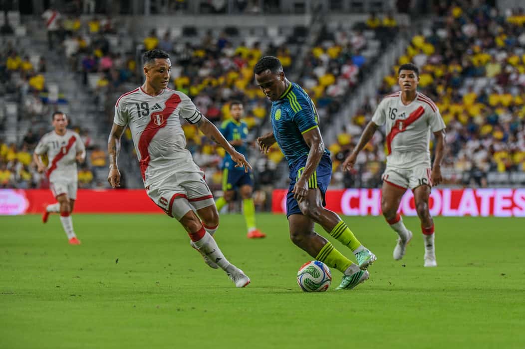 Un jugador de Colombia, con la camiseta azul, encara ante la marca de dos futbolistas de Perú en el Nu Stadium de Miami, con tribunas amarillas de fondo (foto: prensa oficial de la Federación Colombiana de Fútbol)