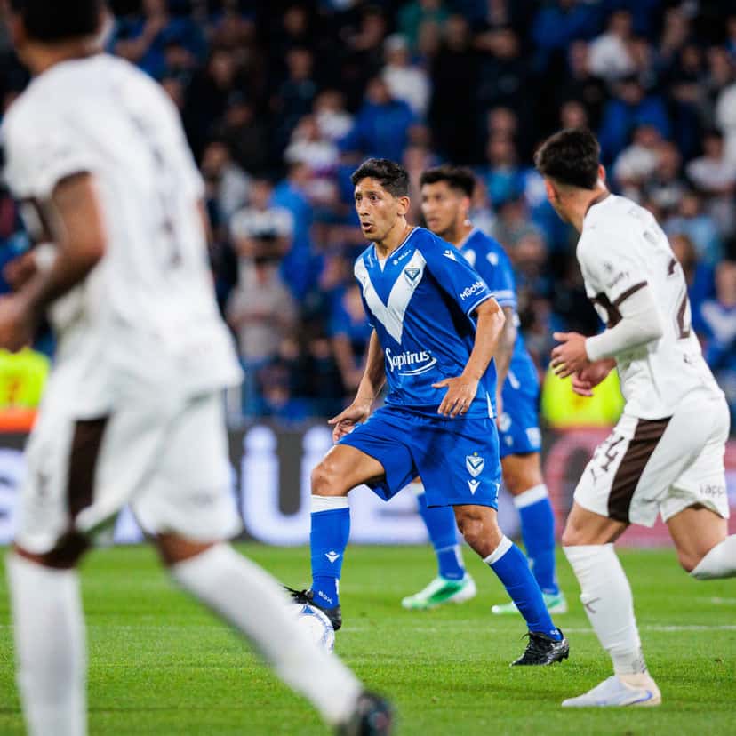 Un jugador de Vélez, con la camiseta azul con la V blanca, conduce la pelota entre dos futbolistas de Platense de camiseta blanca, con la tribuna local de fondo, en el estadio José Amalfitani el lunes 5 de octubre (foto: Hernán Mauricio, prensa oficial de Vélez)
