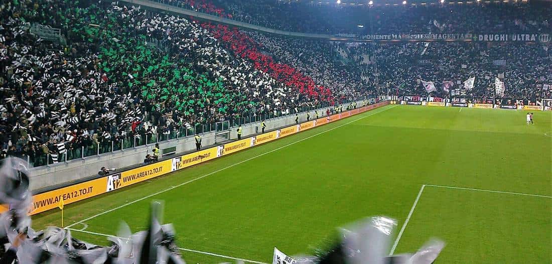 Las tribunas del estadio de Juventus, en Turín, llenas de hinchas con banderas blancas y negras, y el campo de juego iluminado; foto de archivo de 2012 (foto: Gabriele Barberis, Wikimedia Commons, licencia CC BY-SA 3.0 IT)