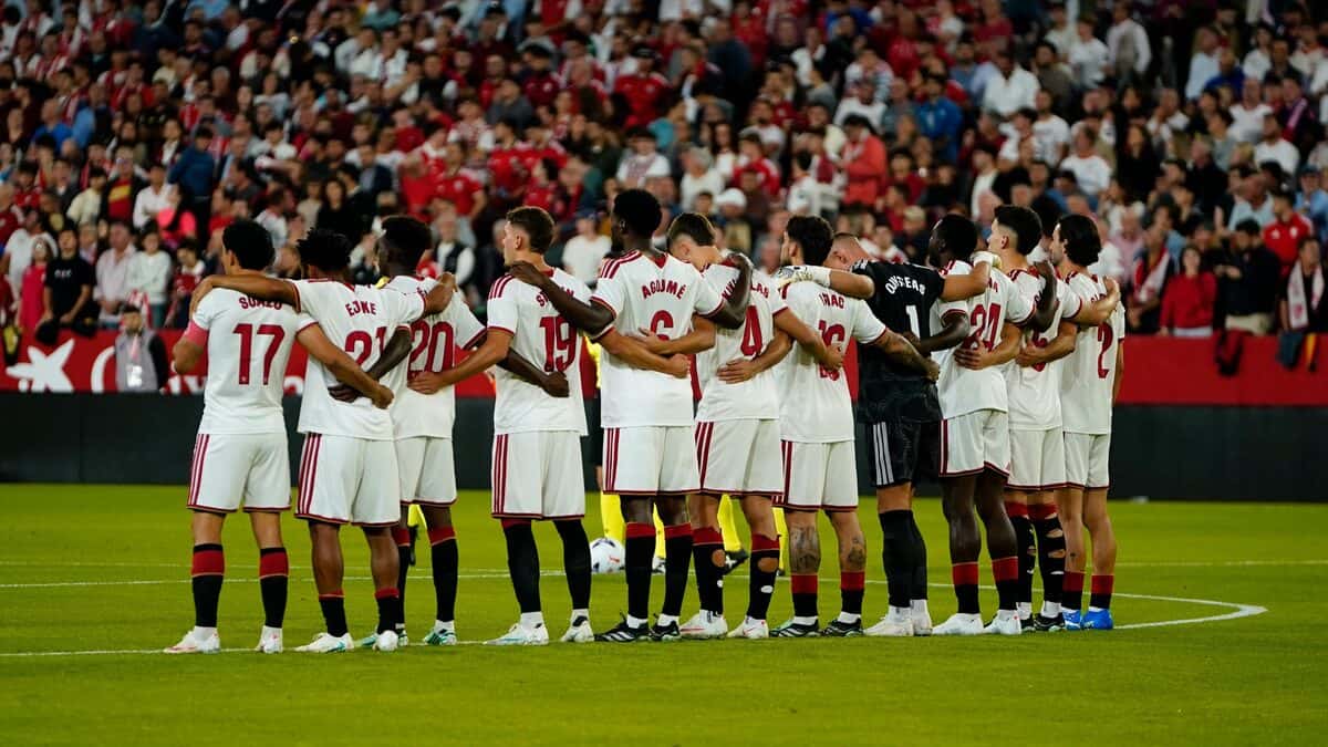 Los jugadores del Sevilla, con la camiseta blanca, abrazados en fila en la mitad de la cancha antes del partido con Osasuna, con la tribuna llena detrás (foto: Sevilla FC)