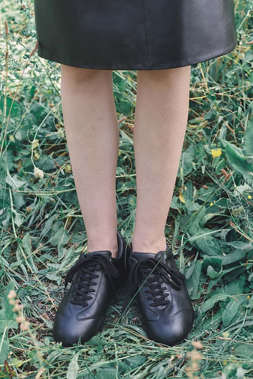 Modelo con la Jil Sander x PUMA Speedcat en cuero negro, parada sobre el pasto con pollera de cuero
