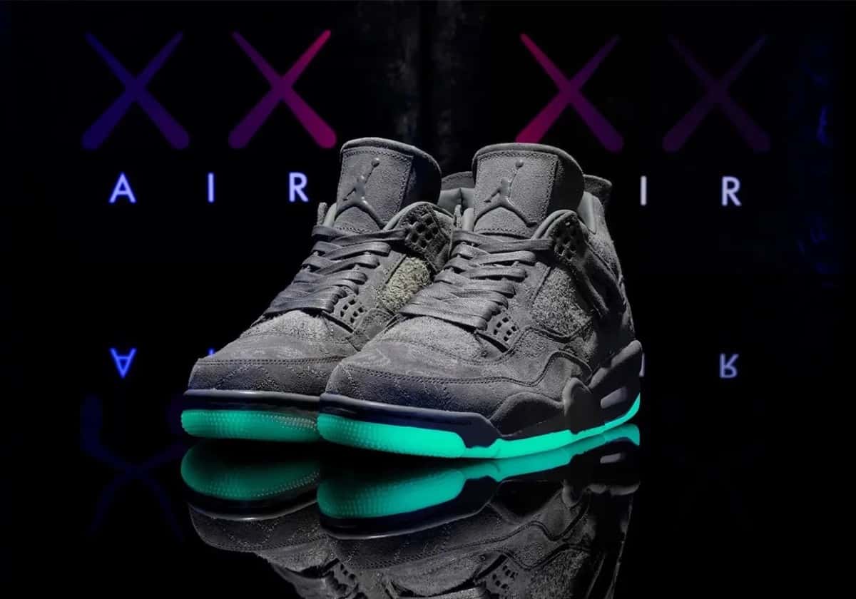 Foto de archivo: KAWS x Air Jordan 4 Cool Grey de 2017 sobre fondo negro, con gamuza gris, suela que brilla en verde agua en la oscuridad y el logo XX AIR en luces de neón detrás