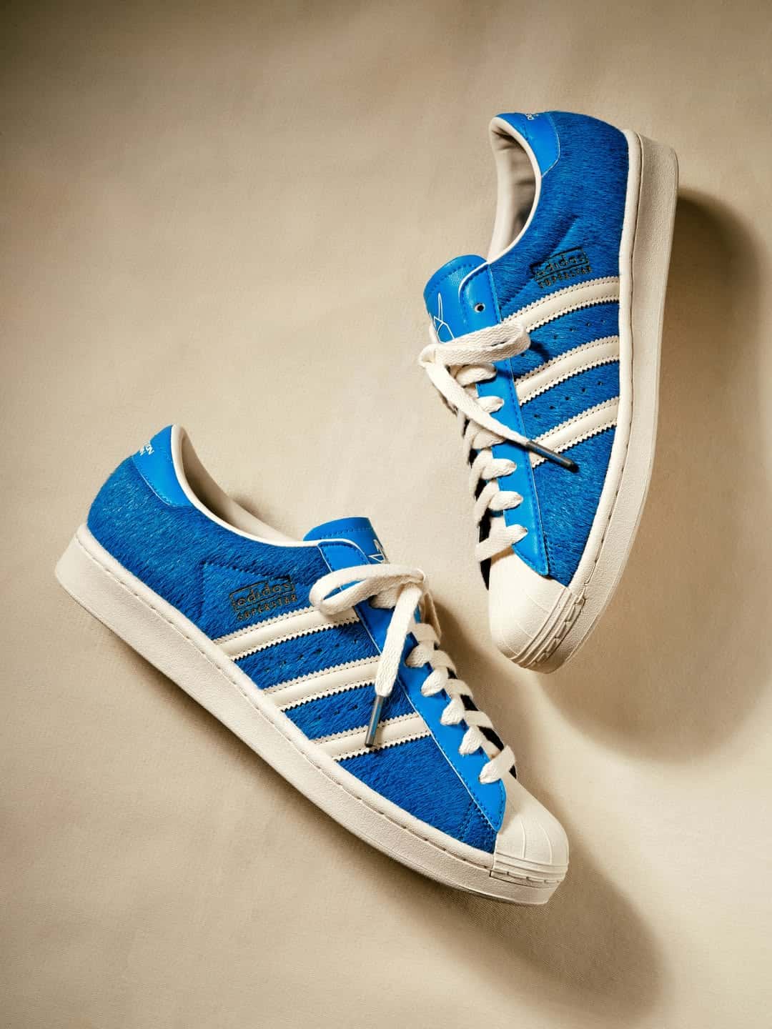 Par de adidas Superstar de Liberaiders sobre fondo beige, con capellada azul de pelo largo, tres tiras y puntera color crema y el logo adidas Superstar en el lateral
