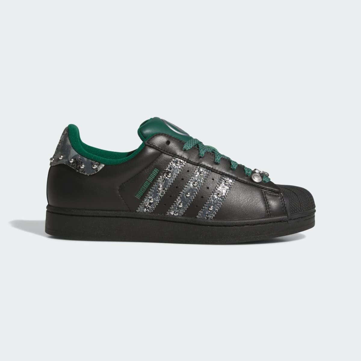 adidas Superstar Doctor Doom de perfil, en cuero negro con tres tiras gris metálico con tachas, cordones y lengüeta verdes y un dije en los ojales