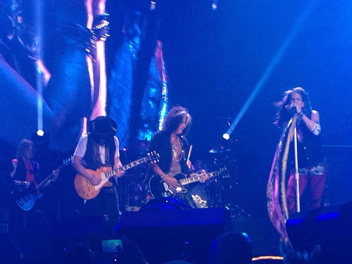 Aerosmith en vivo en Grand Rapids, Estados Unidos, con tres guitarristas a la izquierda y Steven Tyler cantando a la derecha, con pañuelos colgando del pie de micrófono, bajo luces azules (foto de archivo, agosto de 2015)