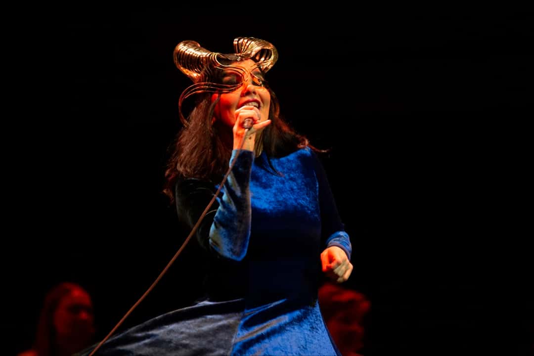 Foto oficial de Björk en el concierto Orkestral del 24 de octubre de 2021: con el vestido de terciopelo azul y el tocado dorado, canta con el micrófono en la mano sobre un fondo oscuro