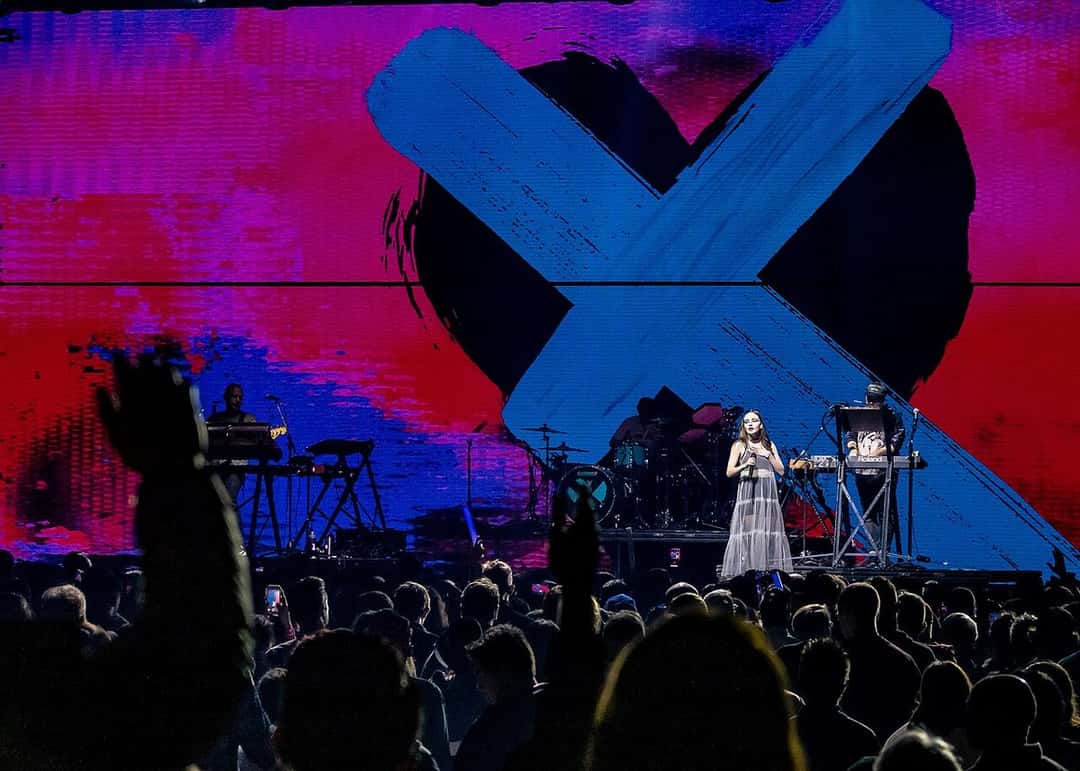 Chvrches en vivo en diciembre de 2018: Lauren Mayberry canta en el centro del escenario, con vestido largo, frente a una pantalla gigante con un corazón negro tachado por una cruz azul sobre fondo rojo y violeta, y el público en primer plano (foto de archivo). Foto: Justin Higuchi, CC BY 2.0, vía Wikimedia Commons