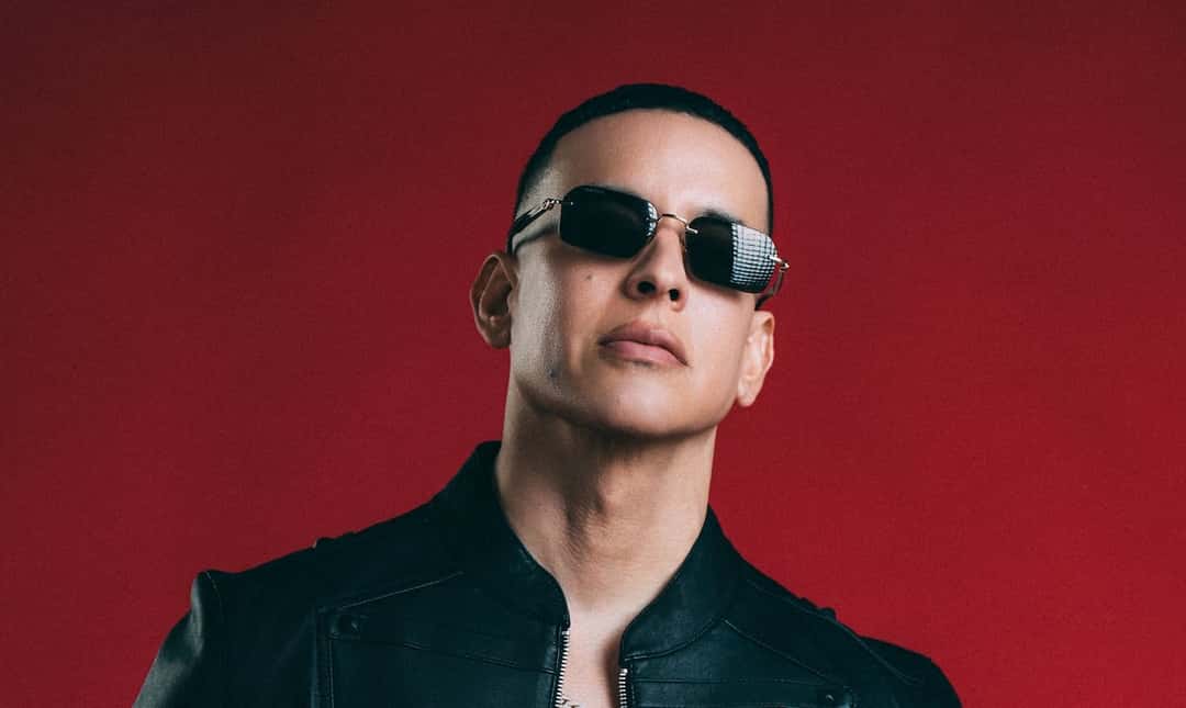 Daddy Yankee con anteojos de sol oscuros y campera negra, frente a un fondo rojo, en una foto de prensa