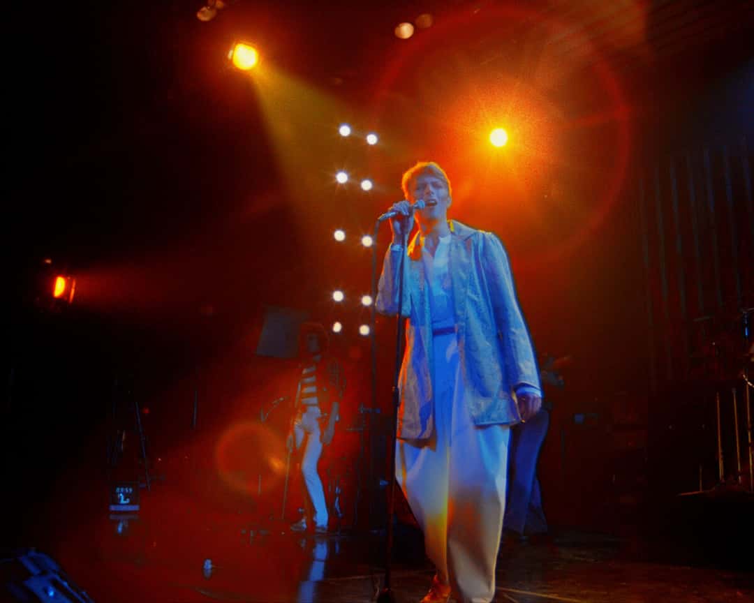 David Bowie, de saco claro y pantalón blanco, canta en el centro del escenario bajo reflectores naranjas, en el show de 1978 (foto de prensa del proyecto «Stage», tomada del material restaurado)