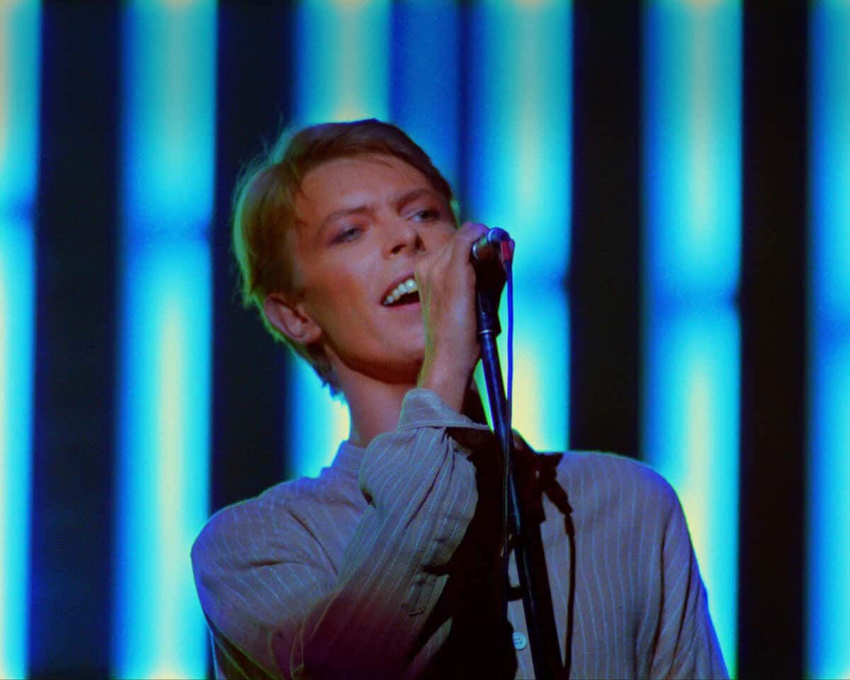 David Bowie canta frente al micrófono, con camisa clara, delante de un fondo de franjas de luz azul, en su show de 1978 en Earls Court (foto de prensa del proyecto «Stage»)