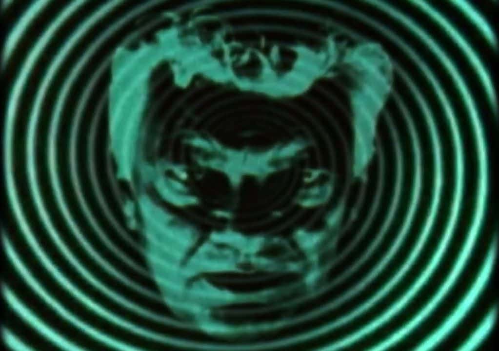 Imagen del teaser oficial «Hypnotism» de Dead Man's Bones: la cara de un hombre en tonos verdes, superpuesta a un espiral hipnótico verde sobre fondo negro