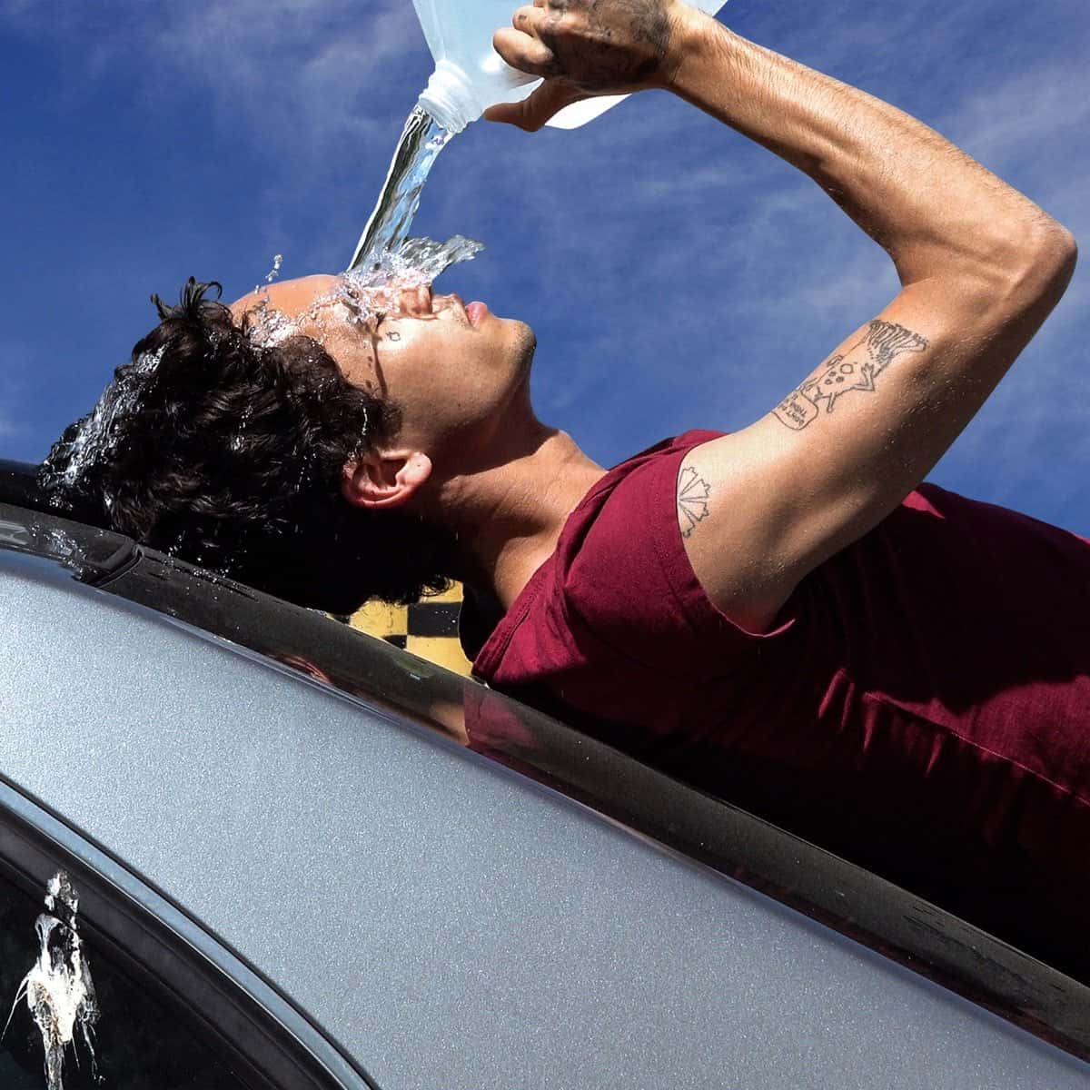 Portada oficial de "How To Quit Smoking" de Dominic Fike: el cantante, con remera bordó, asomado por el techo de un auto plateado, se tira agua de un bidón sobre la cara bajo un cielo azul